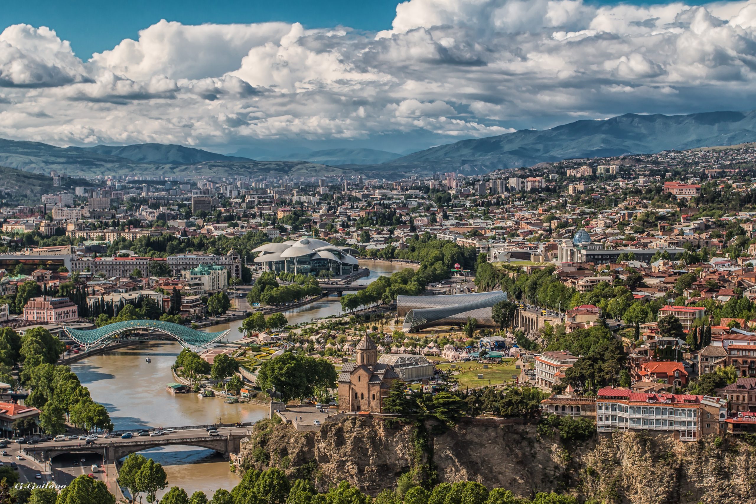 La città di Tbilisi, in Georgia, ripresa dall'alto. C'è un fiume che l'attraversa, si vedono i ponti, le macchine e i tetti delle case. In lontananza ci sono delle montagne e delle nuvole compatte